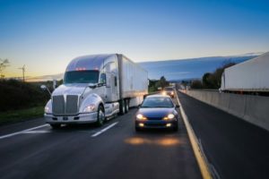 Semi truck and car traveling on an Indianapolis divided highway, traveling the same direction at dusk with their headlights on.